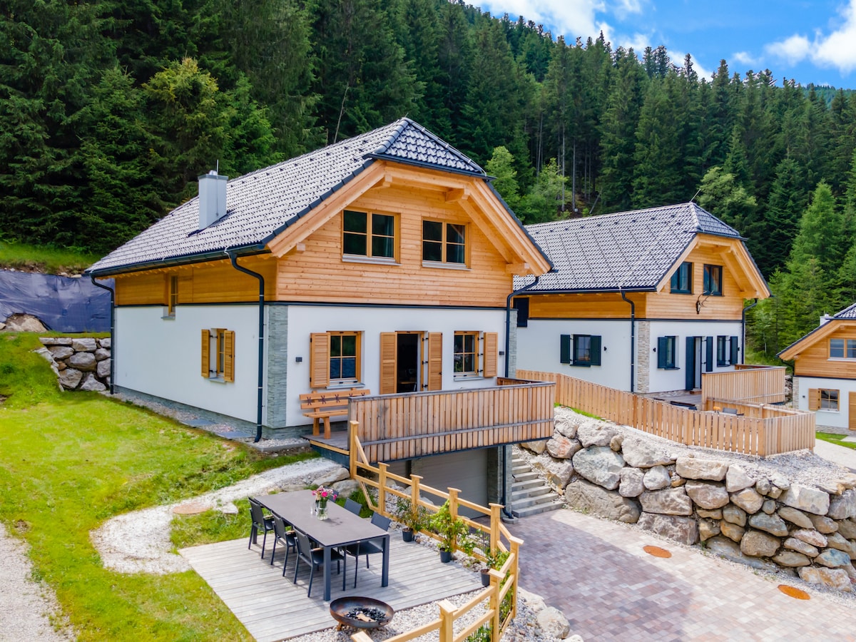 The image captures a pair of chalets featuring a traditional Alpine design, surrounded by lush greenery. A spacious wooden deck with seating is visible in front of one chalet, while a neatly arranged outdoor dining table is positioned nearby. Stone pathways and landscaping enhance the tranquil setting.