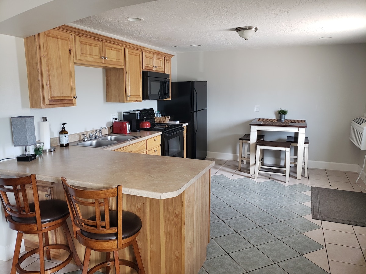 A functional kitchen area is highlighted by wooden cabinetry and a spacious counter with two bar stools. The kitchen includes modern appliances, such as a black refrigerator and microwave. A small dining table set against the wall offers additional seating, accented by neutral decor.