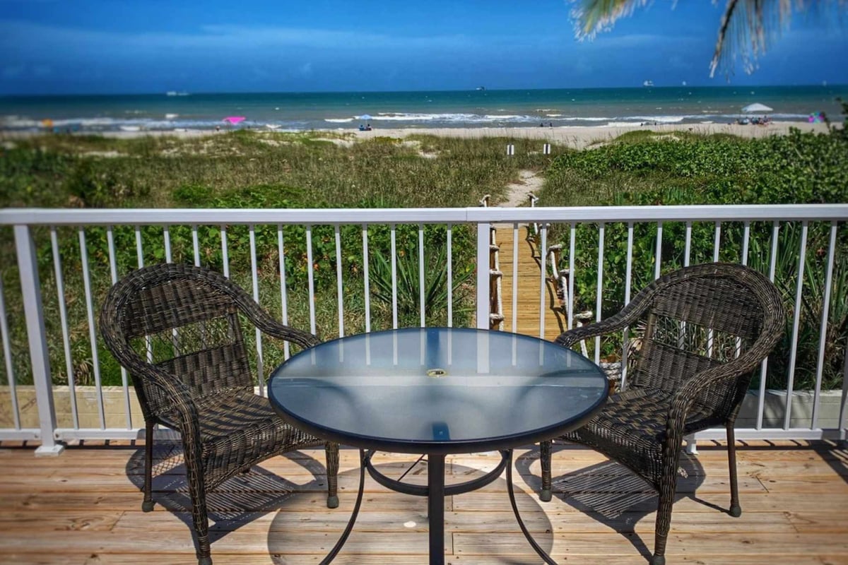 A deck with a circular table and two matching chairs overlooks the ocean. The view includes sandy shores and green coastal vegetation, with beachgoers visible in the distance. The sky is clear, providing a serene atmosphere for relaxation.