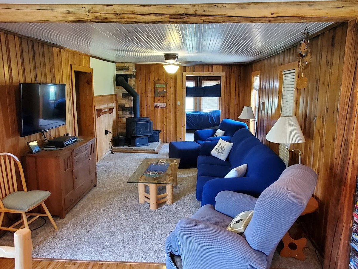 A cozy living room features wooden walls and a plush blue sectional sofa, complemented by a recliner and a wooden rocking chair. A coffee table rests on soft carpeting, with a television mounted on one wall and a rustic fireplace at the opposite end.