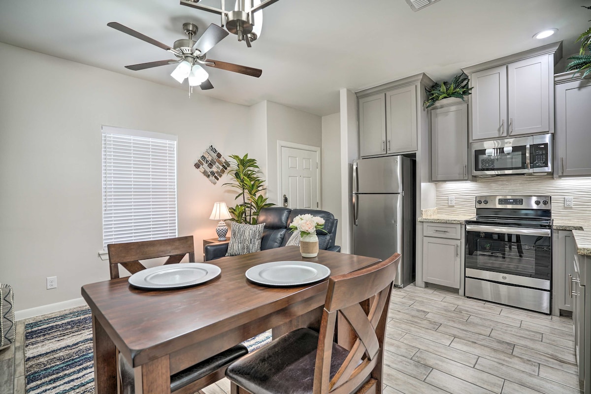 A modern dining area features a wooden table set for two, complemented by comfortable chairs. In the background, an open kitchen is equipped with stainless steel appliances and sleek cabinetry. Natural light filters through a window, highlighting the homey ambiance.