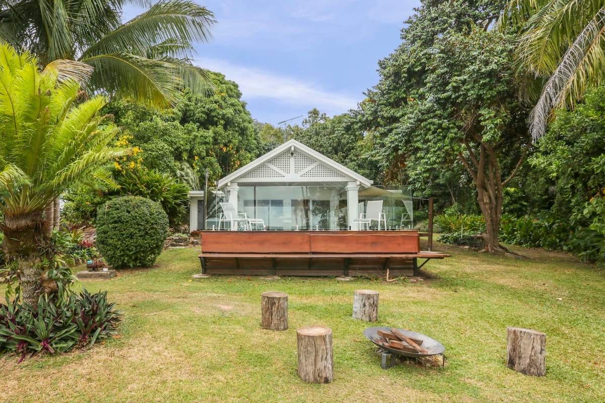 A charming retreat is surrounded by lush greenery, featuring a wooden seating area with log stools and a fire pit. The spacious house, with expansive glass doors, provides a view of the serene outdoor landscape and is framed by tropical plants.