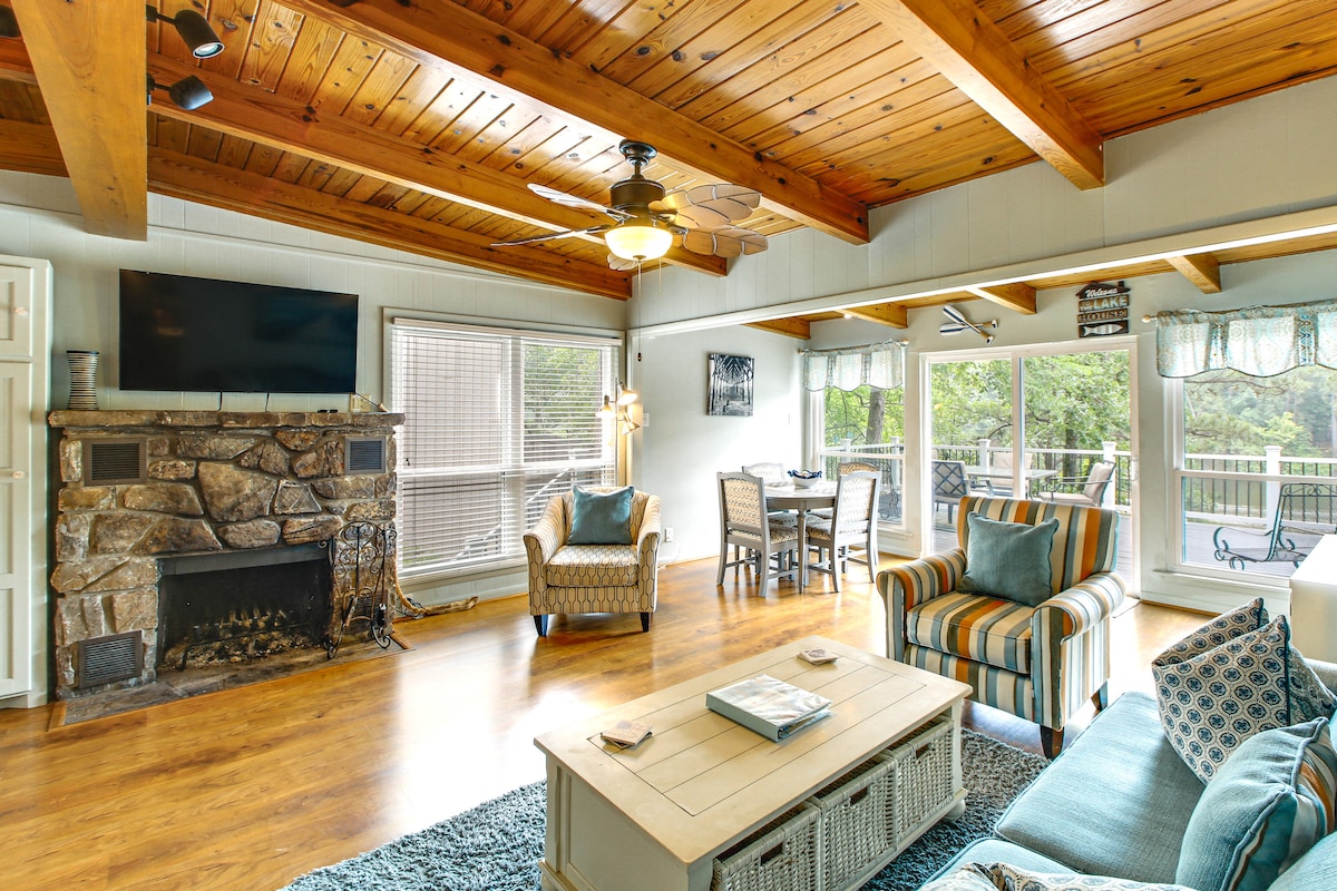 A bright living area features wooden beams across the ceiling and a stone fireplace. Comfortable seating arrangements include patterned chairs and a couch with blue accents. Large windows provide a view of the outdoor space, while a dining table is positioned nearby, completing the welcoming environment.