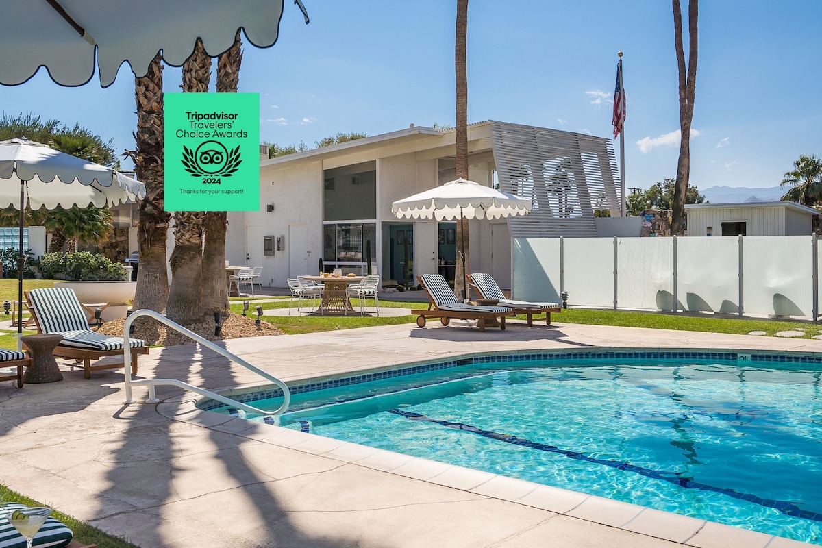 A serene pool area is framed by lounge chairs and umbrellas, inviting relaxation under the sun. The modern hotel building features large glass doors, offering views of the lush greenery and mountains in the background, complemented by the warm hues of the spacious patio.
