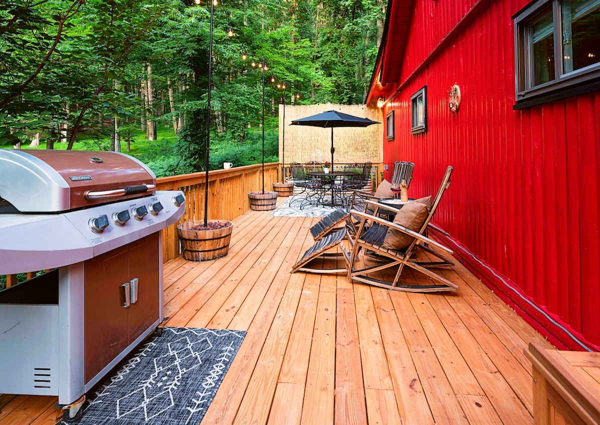 A spacious wooden deck features a gas grill and several seating options, including custom Adirondack chairs. An umbrella-shaded dining table provides a space for meals, while string lights add a warm touch. Lush greenery surrounds the area, enhancing the outdoor experience.