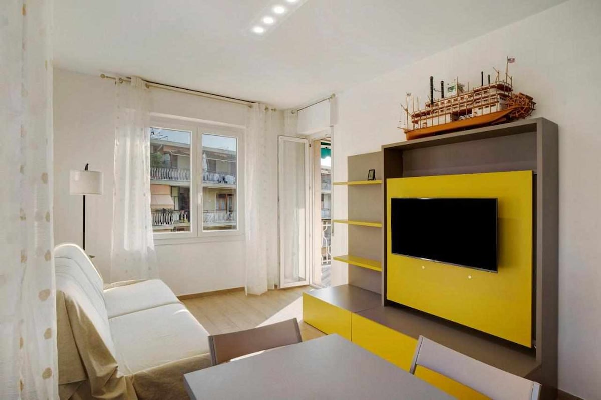 A bright living area is furnished with a white sofa and a modern TV mounted on a yellow entertainment unit. A dining table with chairs is placed near the window, which allows natural light to fill the room. Light curtains frame the window, enhancing the space.