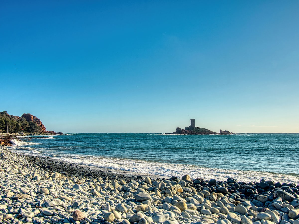 A scenic view of a rocky beach is depicted, with smooth stones lining the shore. Gentle waves roll in towards the beach, and a distant island features a stone tower under a clear blue sky, creating a tranquil coastal atmosphere.