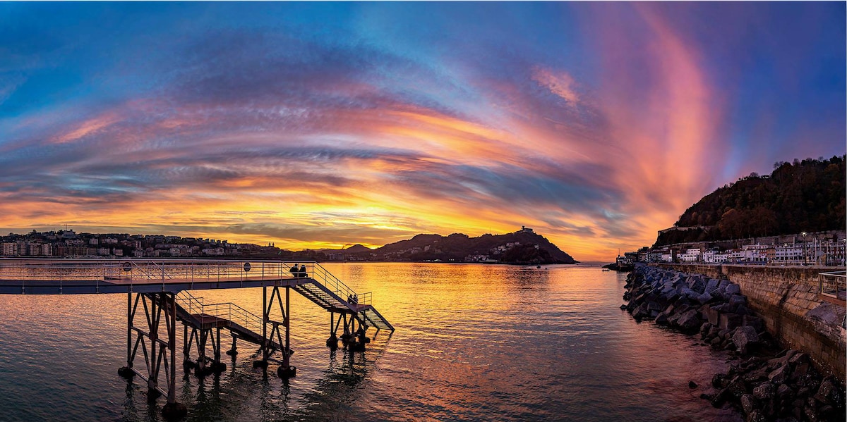 A panoramic view captures the serene coastline at sunset, with vibrant colors reflecting on the water. A wooden dock extends into the sea, framed by silhouettes of surrounding hills and city structures along the shore.