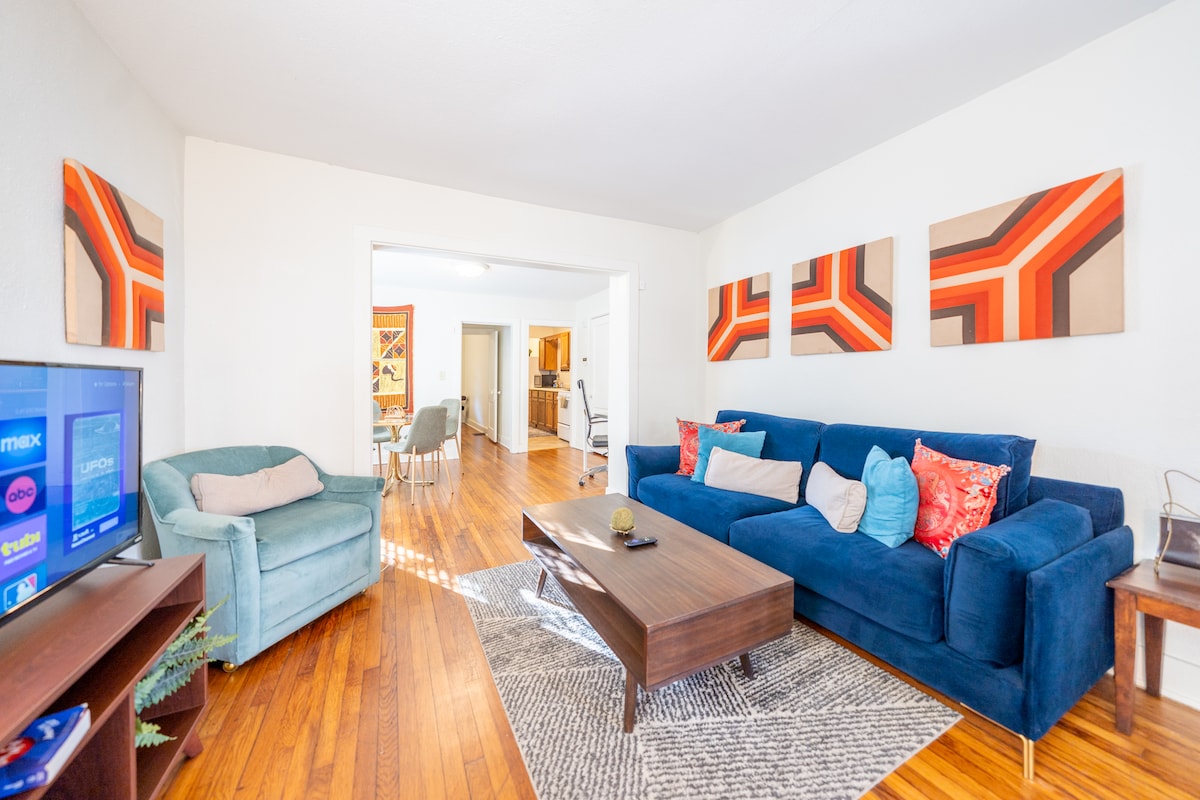 The living area features a spacious blue couch adorned with vibrant throw pillows, complemented by a cozy armchair. A wooden coffee table sits at the center, while modern artwork adorns the walls, enhancing the environment. Natural light is welcomed through the nearby doorway.