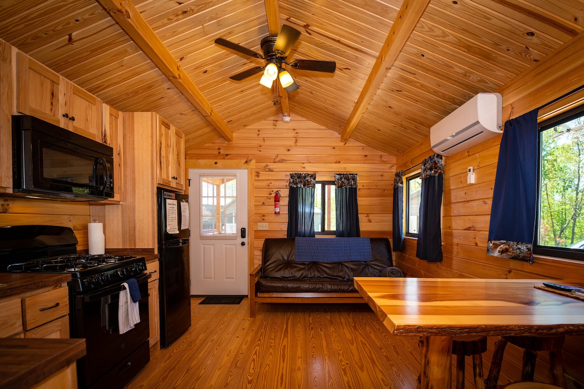 The interior space features natural wood paneling and a high ceiling. A comfortable futon sits to one side, complemented by a dining table. A kitchen area includes modern appliances such as a microwave and gas range. Windows allow natural light to fill the room.