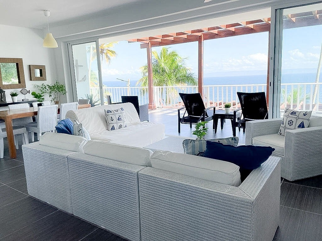 A spacious living area features a light-colored sofa with nautical-themed cushions, facing large glass doors that open to a breathtaking ocean view. A wooden dining table and chairs are positioned nearby, with tropical plants adding a touch of greenery.