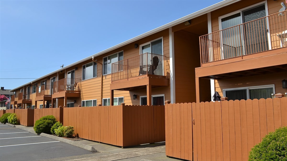 A row of ground floor condominiums features private patios with wooden railings. Each unit includes sliding glass doors, allowing natural light to enter. Lush greenery is present in front of the fencing, providing a welcoming outdoor space. Two designated parking spaces are visible in the foreground.