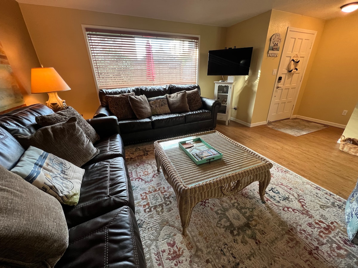 A comfortable living room features dark leather sofas arranged around a patterned area rug. A coffee table sits in the center, adorned with decor. Natural light filters through a large window, with a view of outdoor space visible. A flat-screen TV is mounted on the wall.