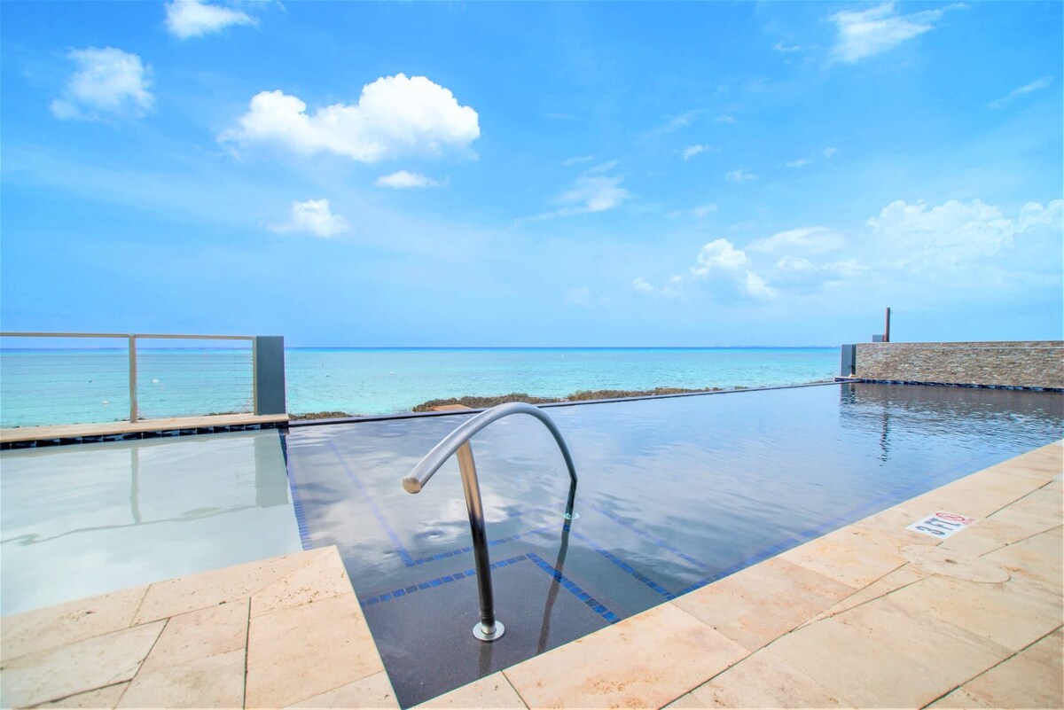 An infinity pool meets the ocean, with a sleek handrail extending into tranquil water. The horizon showcases a blend of blue seas and cloudy skies, while a stone wall provides a modern backdrop, enhancing the serene coastal atmosphere.