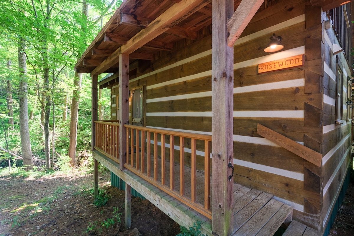 The exterior of the Rosewood cabin is showcased, featuring natural wood siding and a welcoming porch. A wooden railing borders the deck, surrounded by lush greenery, providing a tranquil connection to nature. A light fixture illuminates the cabin's name sign, showcasing its charm.