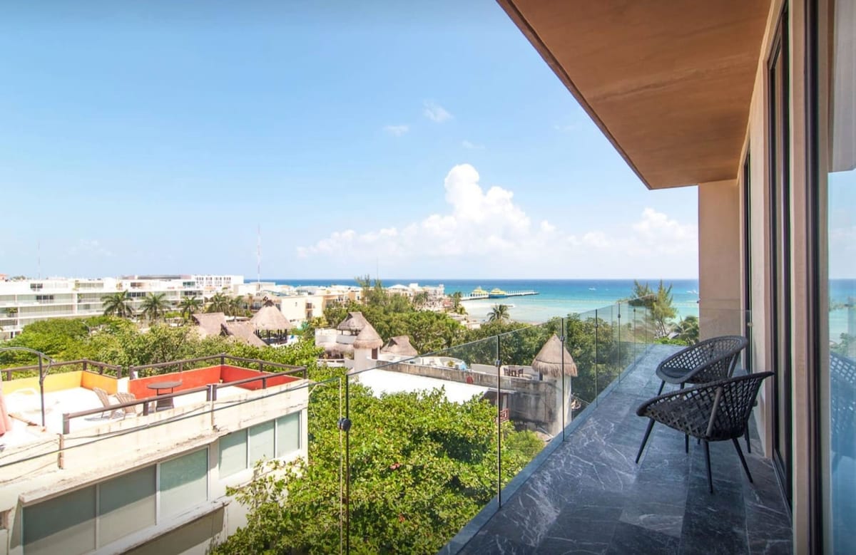 A balcony offers breathtaking views of the Caribbean Sea and surrounding greenery. Two modern chairs are positioned on the balcony, providing a perfect spot to relax and enjoy the scenery. The clear sky and distant beach complement the inviting atmosphere.