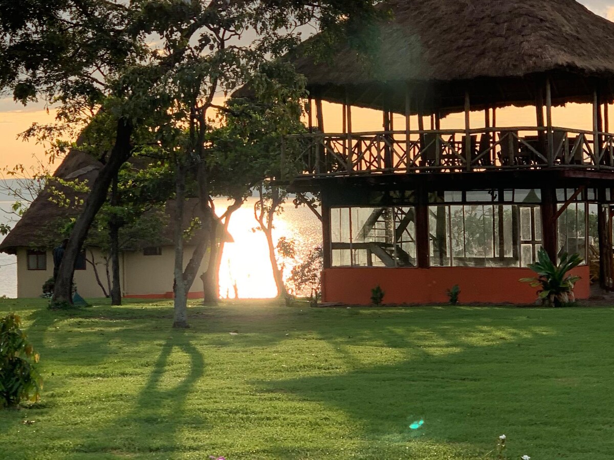 A grassy area is seen with the silhouette of trees casting shadows. Two distinct structures with thatched roofs are visible, along with an open balcony area facing towards a shimmering body of water in the background under a soft sunset glow.