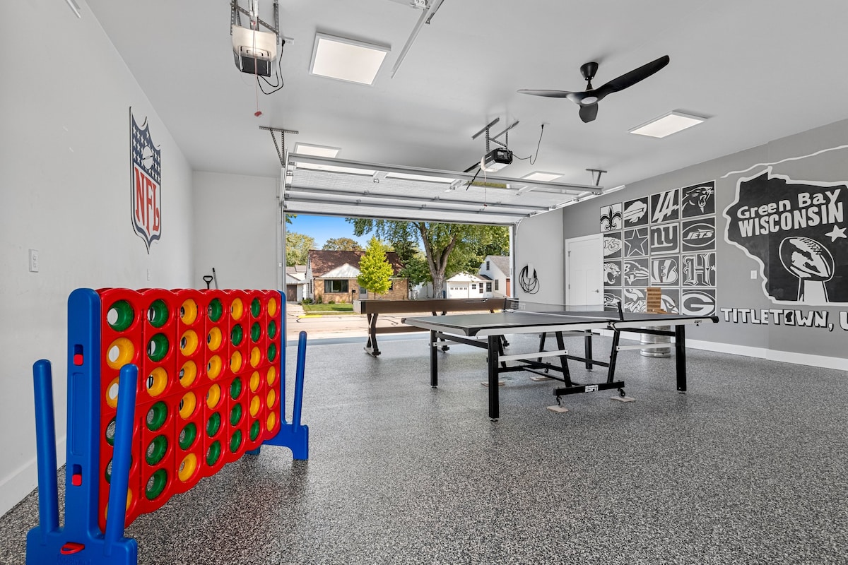 A heated garage space is equipped for entertainment, featuring a ping pong table and a Connect Four game. The walls display local sports team pride, and ample overhead lighting enhances the airy atmosphere. A garage door provides natural light and exterior views.