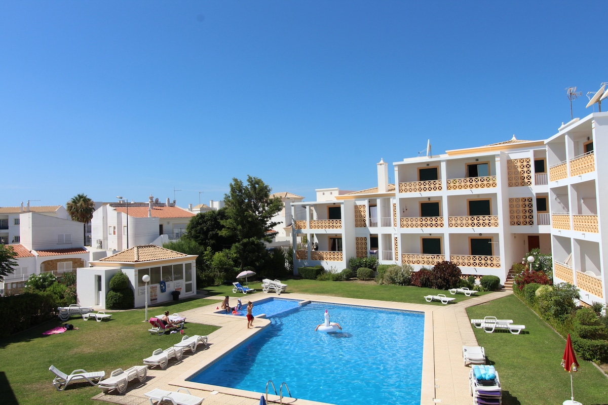 A view of the outdoor pool area within a well-maintained condominium. Lounge chairs are arranged around the pool, and guests can be seen enjoying the water. Lush greenery and well-kept gardens surround the pool, and buildings can be seen in the background, adding to the spacious ambiance.