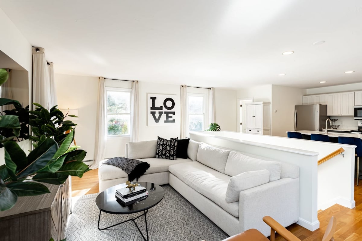 A bright living area features a spacious light-colored sofa with black and white accents, positioned near a round coffee table. Natural light pours through large windows, illuminating the soft décor. A large wall art piece spells 'LOVE' above the sofa, enhancing the welcoming ambiance.