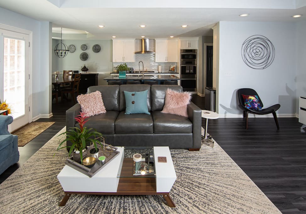 The open-concept living space features a dark leather sofa adorned with colorful cushions, centered on a modern coffee table. Natural light streams through sliding glass doors, highlighting the sleek kitchen and dining area in the background. Subtle decor elements add an inviting touch.