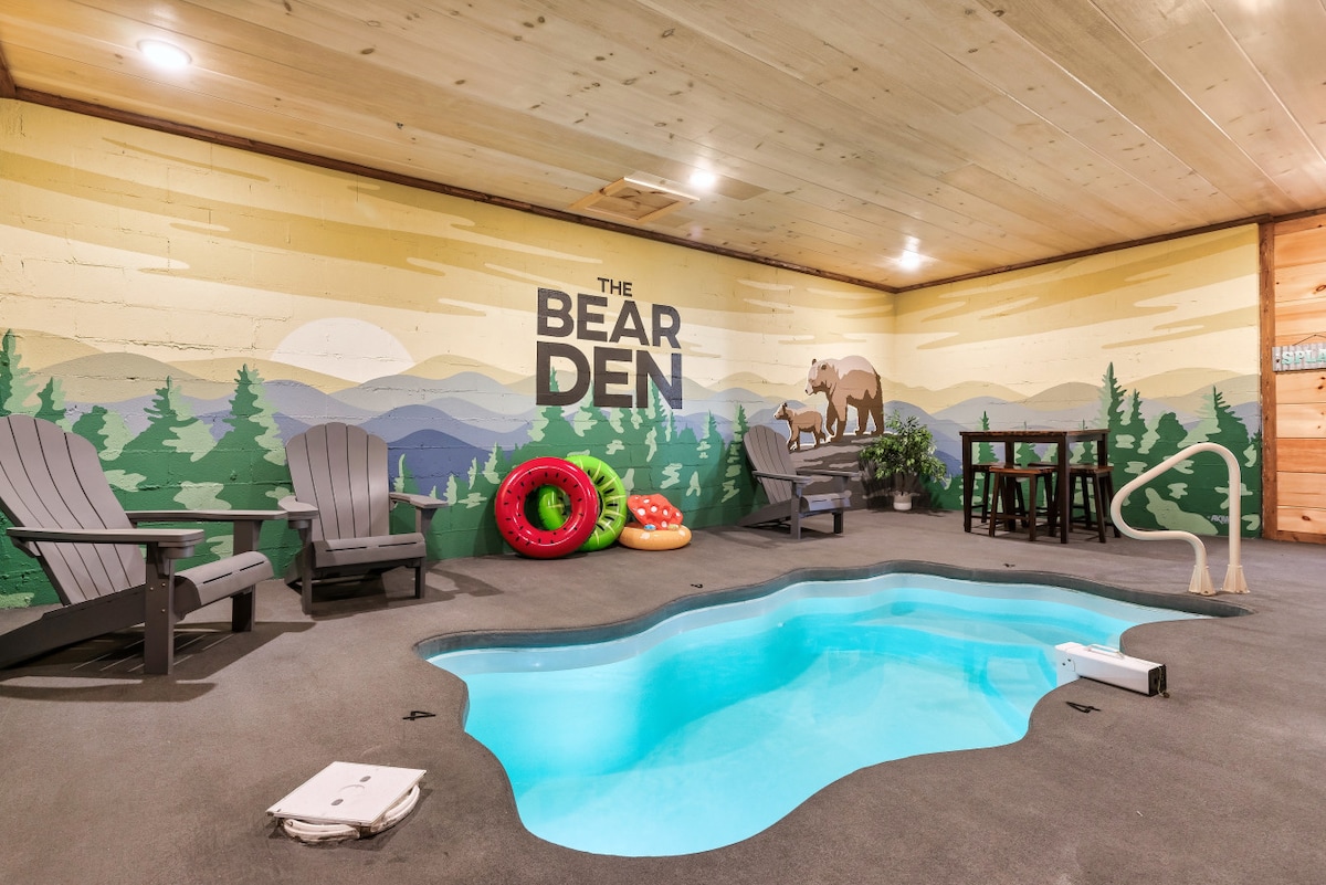An indoor pool area features a turquoise pool surrounded by a soft gray floor. Two adirondack chairs are placed beside the pool, and colorful pool floats rest against the wall. A mural of a bear and mountains adds a festive touch to the space.