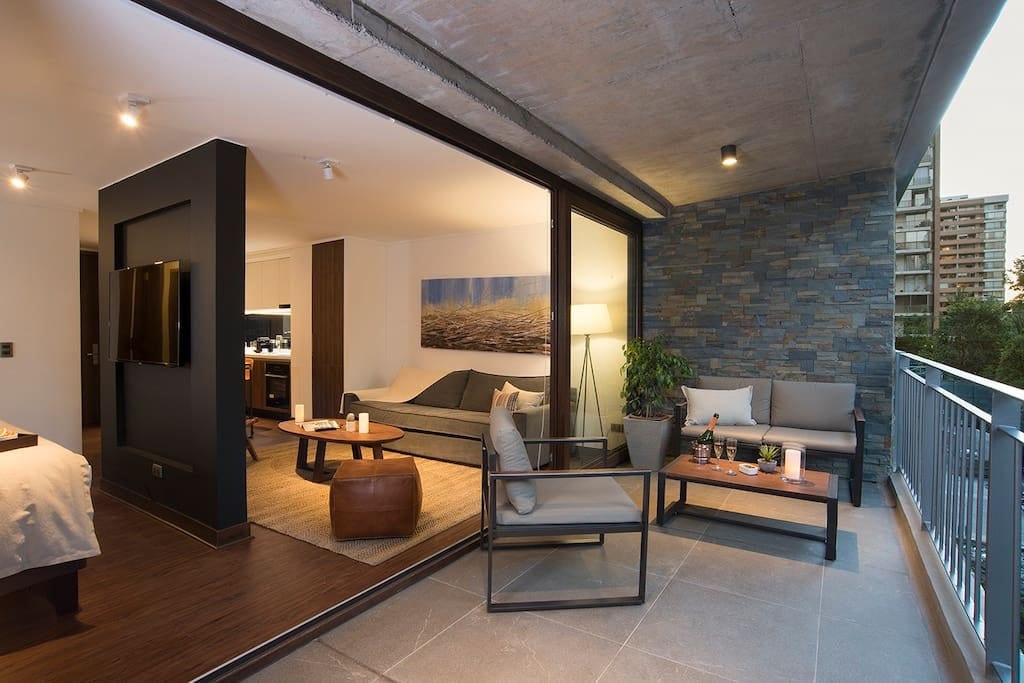 An outdoor terrace features modern seating arrangements, including a sofa and armchairs, with a round table set for beverages. Large glass doors lead into a bright interior space decorated in neutral tones, complemented by a stone accent wall and natural light filtering in.