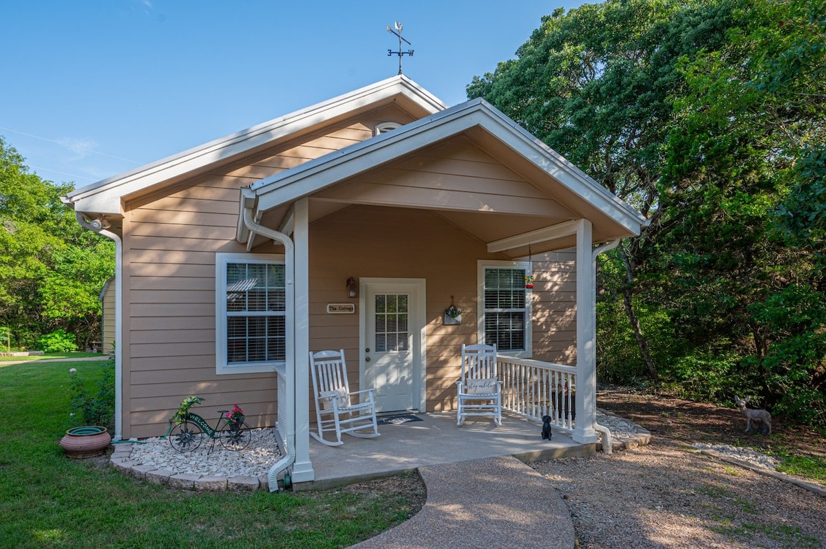 A charming cottage exterior is shown, featuring a welcoming entryway with a covered porch. Two rocking chairs are positioned beside a small side table. The surrounding landscape is lush with greenery, creating a serene entrance to the home.