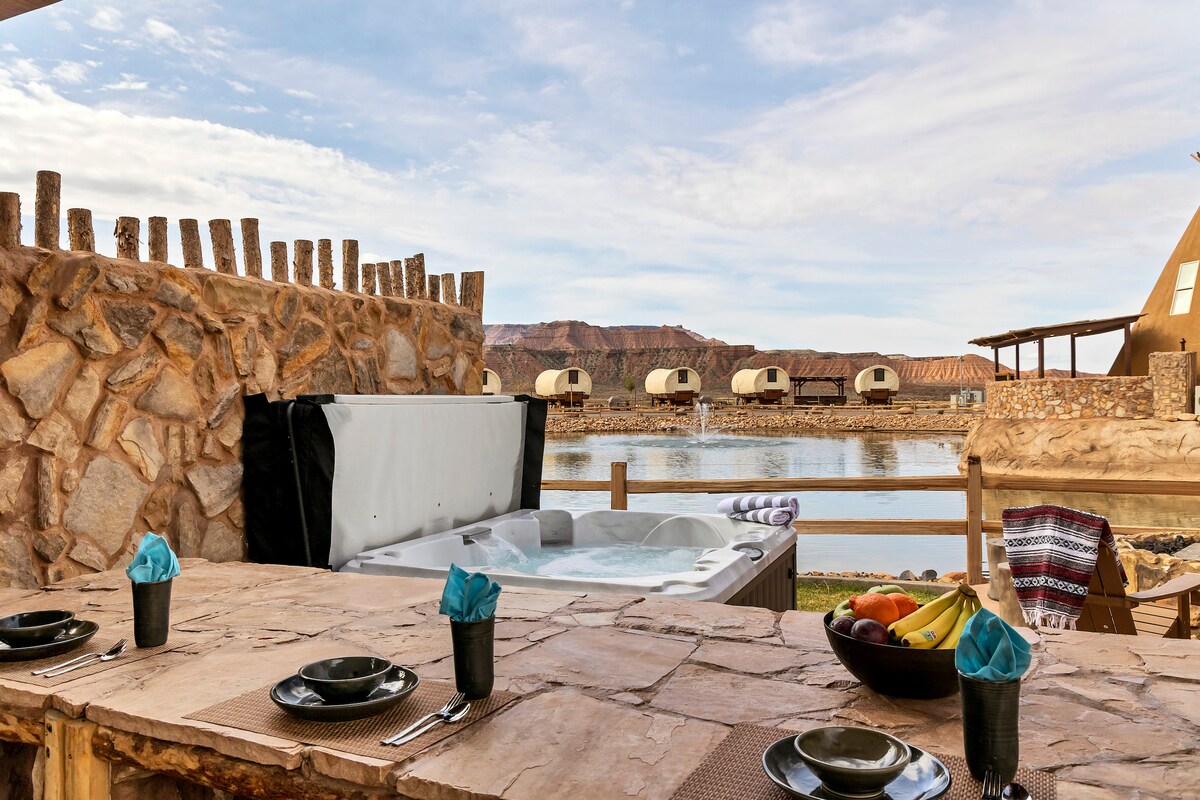 A private hot tub with bubbles is set on a stone patio overlooking a tranquil pond. A wooden bar area features plates and napkins arranged neatly, accompanied by a bowl of fresh fruits. Tipis are visible in the background against a scenic landscape.