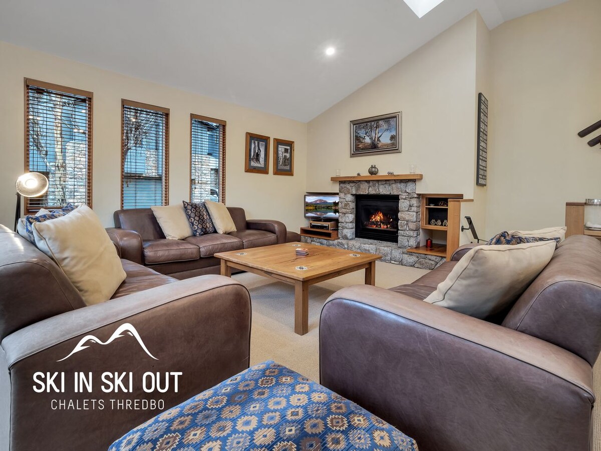 A spacious lounge area features comfortable leather seating arranged around a central wooden coffee table. A stone fireplace adds warmth to the room, with natural light streaming in through several windows dressed with wooden blinds.