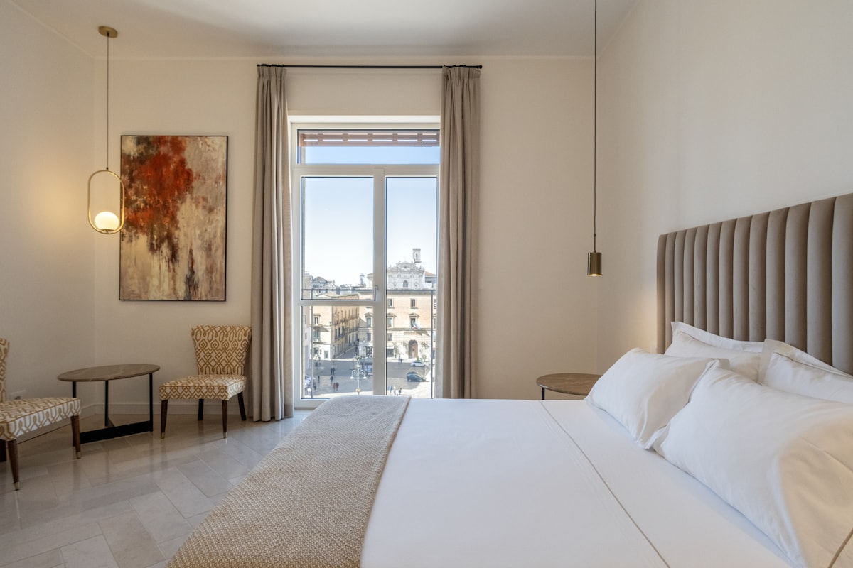 A spacious guest room features a king-size bed dressed in high-quality linens. Two elegant chairs are positioned beside a small table. Natural light fills the room through a large window, which offers a view of the historic square outside, framed by soft drapes.