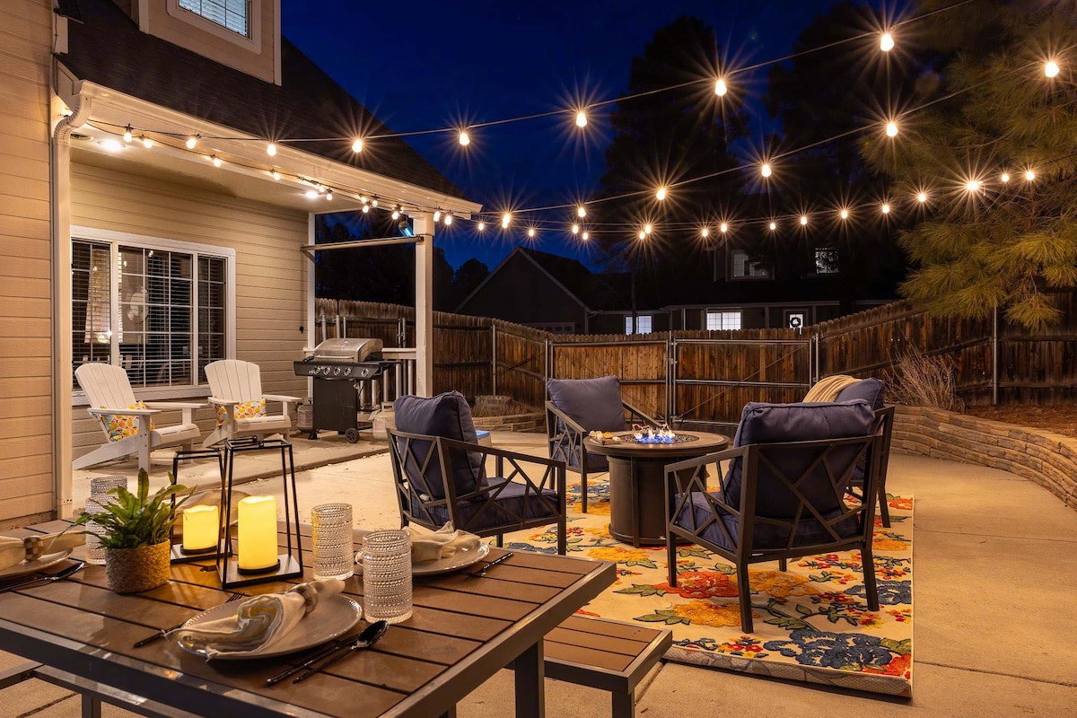 An outdoor seating area is illuminated by string lights and features comfortable chairs surrounding a fire pit. A dining table is decorated with dinnerware and a plant. A grill is visible in the background, providing options for al fresco cooking.