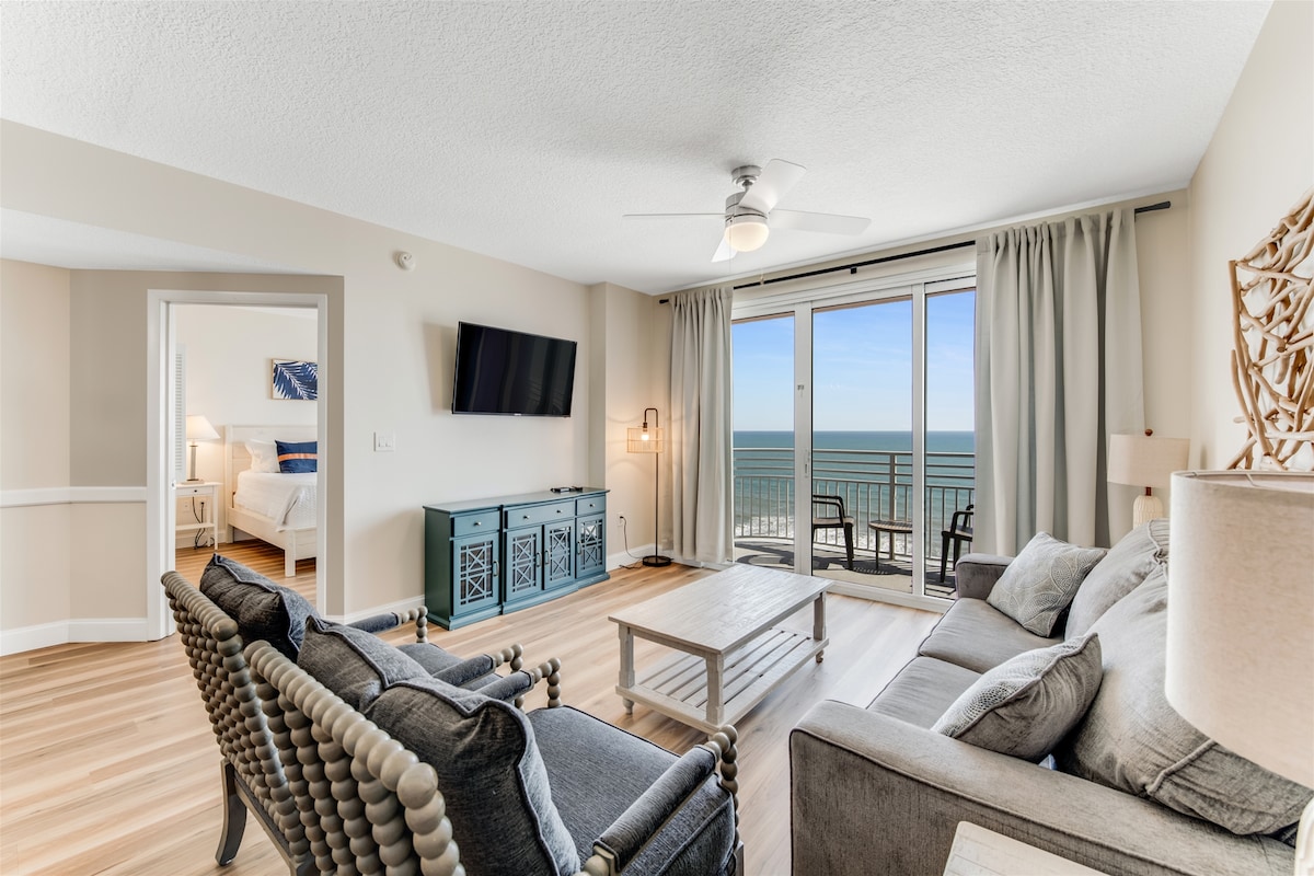 A spacious living area features light-toned wood flooring and large sliding glass doors that frame a direct ocean view. Comfortable seating is arranged around a wooden coffee table, while soft lighting and neutral décor create a relaxing environment.