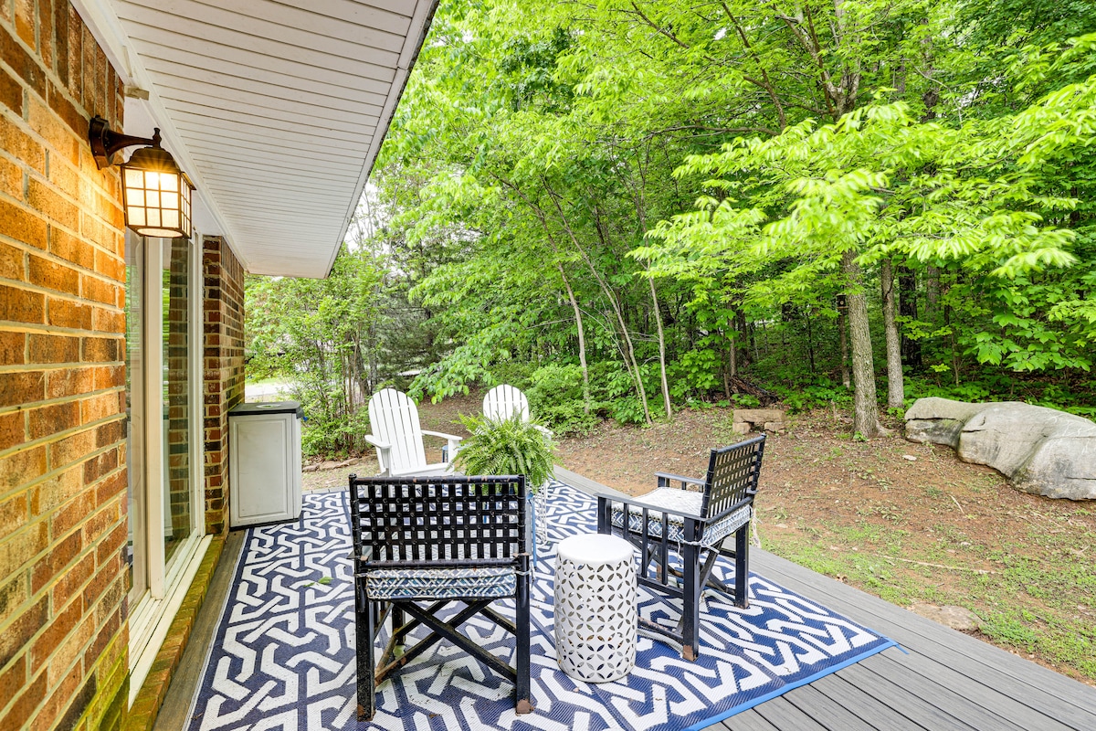 A welcoming outdoor seating area showcases two black chairs and a small circular table on a patterned rug. Lush greenery frames the space, providing a serene environment. Natural stone elements are visible among the foliage, contributing to the tranquil atmosphere.