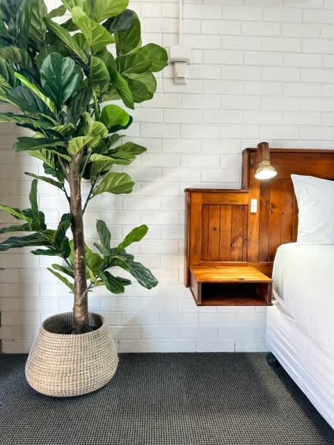 A spacious bed is positioned near a white brick wall, with a wooden bedside table featuring a built-in lamp. A decorative potted plant enhances the area, contributing to a calming atmosphere.