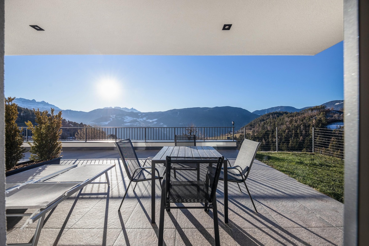 An outdoor terrace is equipped with a dining table and chairs, offering a serene view of the surrounding mountains. Lounge chairs are positioned for relaxation, while sunlight bathes the area, enhancing the pleasant atmosphere of the space.