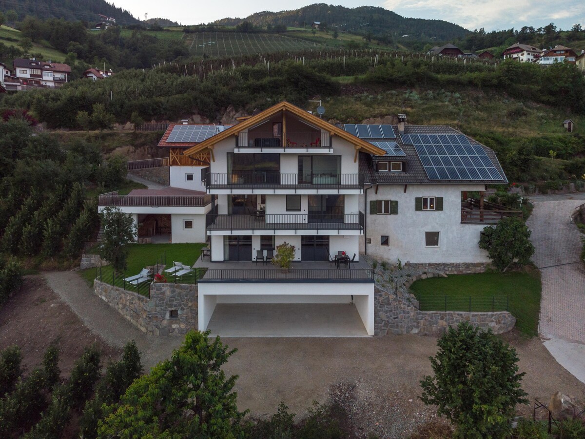 The exterior of the apartment showcases a modern three-story building with a large balcony and two levels of outdoor spaces. Solar panels are visible on the roof. Surrounding greenery includes trees and open fields, contributing to the scenic hillside setting.