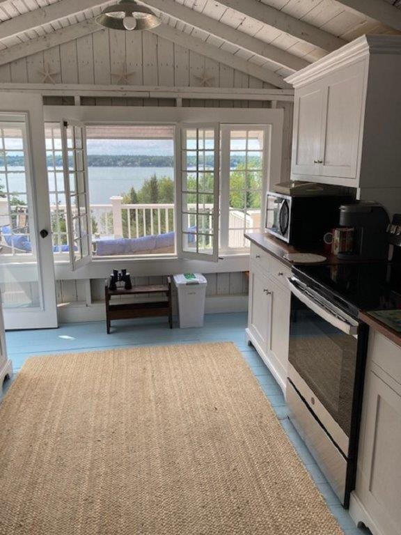A modern kitchen is highlighted with white cabinetry and stainless steel appliances. Large windows frame scenic views of the water beyond, allowing natural light to fill the space. A neutral-colored area rug adds warmth to the blue floor, while a small table sits near the entrance.