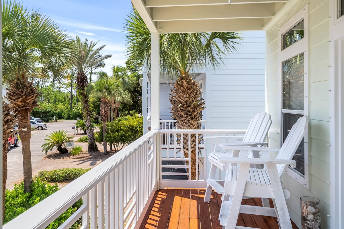 A spacious covered balcony featuring two white adirondack chairs is surrounded by lush palm trees. The wooden flooring complements the light-colored exterior of the townhome, while a view of the community landscape is visible in the background.