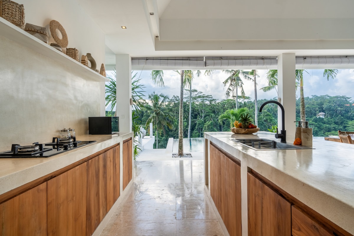 The kitchen features modern appliances, including a gas stove and double sinks set in natural wood cabinetry. Large windows provide a view of the lush jungle, while the open layout connects to the outdoor living area, enhancing the sense of space and tranquility.
