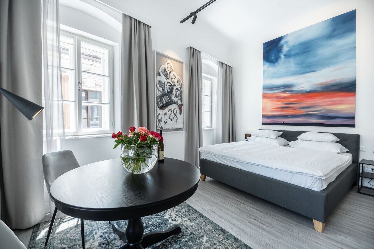 A spacious studio is centered around a king-size bed with crisp white bedding. A round black dining table is accompanied by a floral arrangement. Large windows adorned with soft gray curtains provide natural light, while an abstract painting in warm tones enhances the wall.