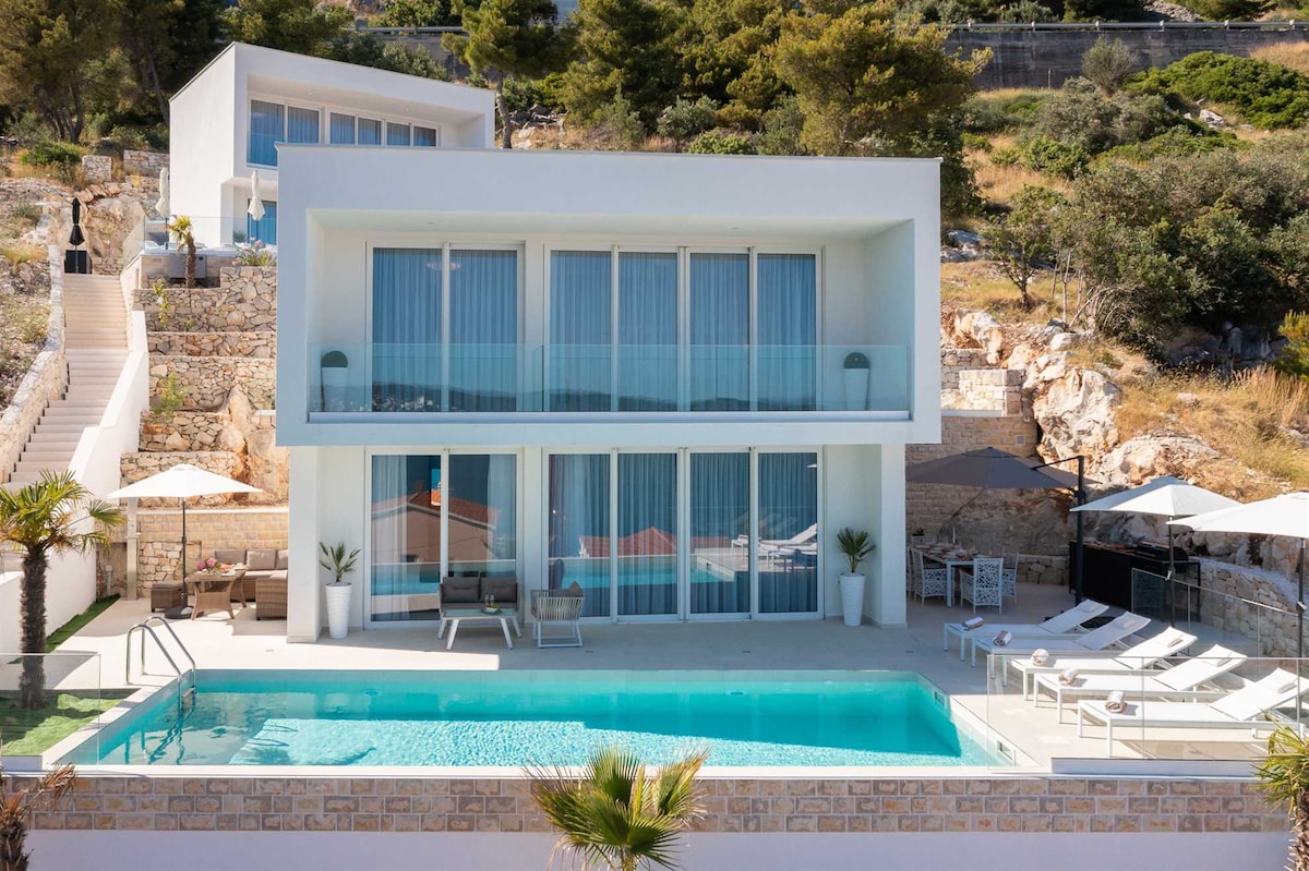 The modern villa features a clean architectural design with large glass windows, allowing natural light to fill the space. An inviting pool area is surrounded by lounge chairs and parasols, complemented by lush greenery and stone accents in the landscape.