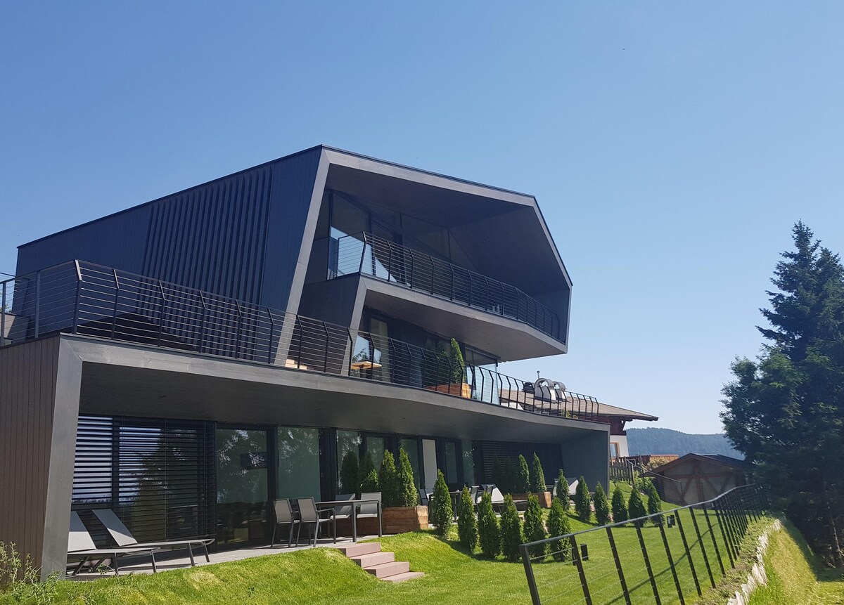 A modern architectural structure features a sleek design with clean lines and multiple levels. The building is surrounded by well-maintained grass and symmetrical shrubbery. A series of loungers are positioned on the terrace, providing a space for relaxation. Large windows reflect the clear blue sky.