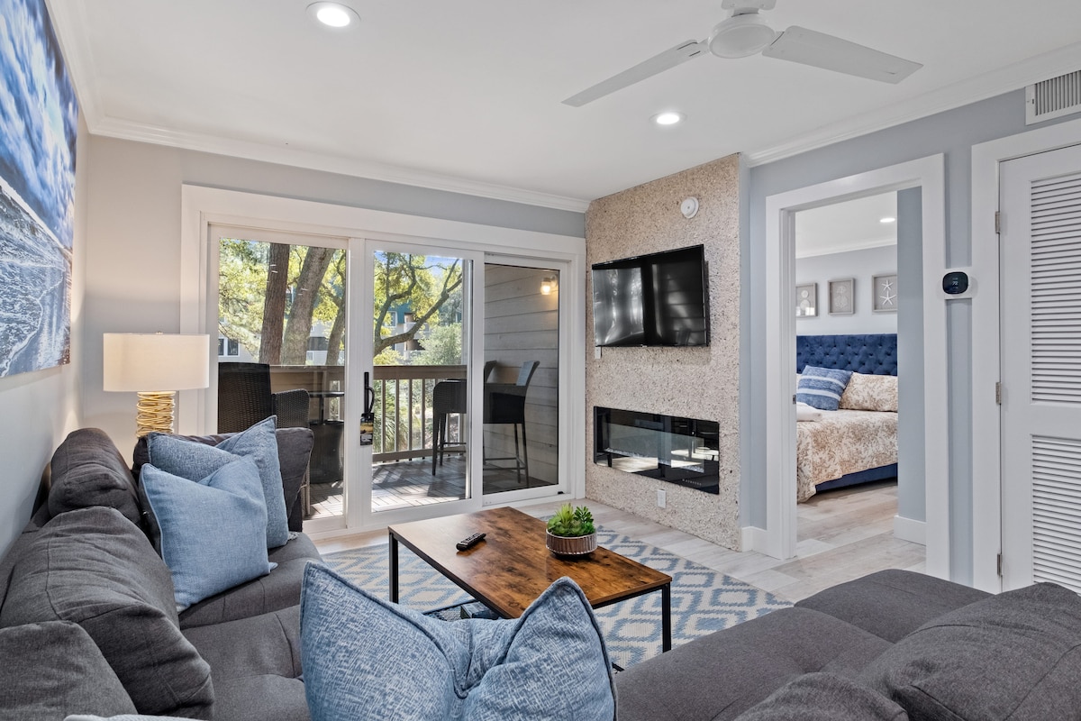 A cozy lounge area is displayed featuring a large sectional sofa adorned with blue accent pillows. Natural light enters through sliding glass doors, which open to a private balcony. A modern wall-mounted TV is positioned above a textured electric fireplace, adding to the room's comfort.