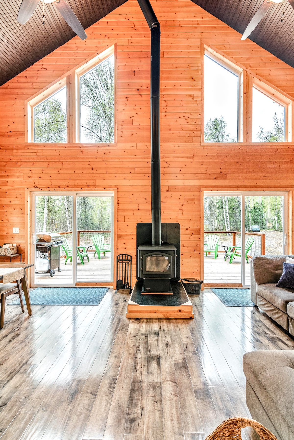 A warm and inviting interior features high ceilings with large windows, allowing natural light to illuminate the wooden walls. A wood stove is positioned centrally, and comfortable seating is arranged nearby, creating a welcoming space for gathering. Views of the outdoor deck are visible through the windows.