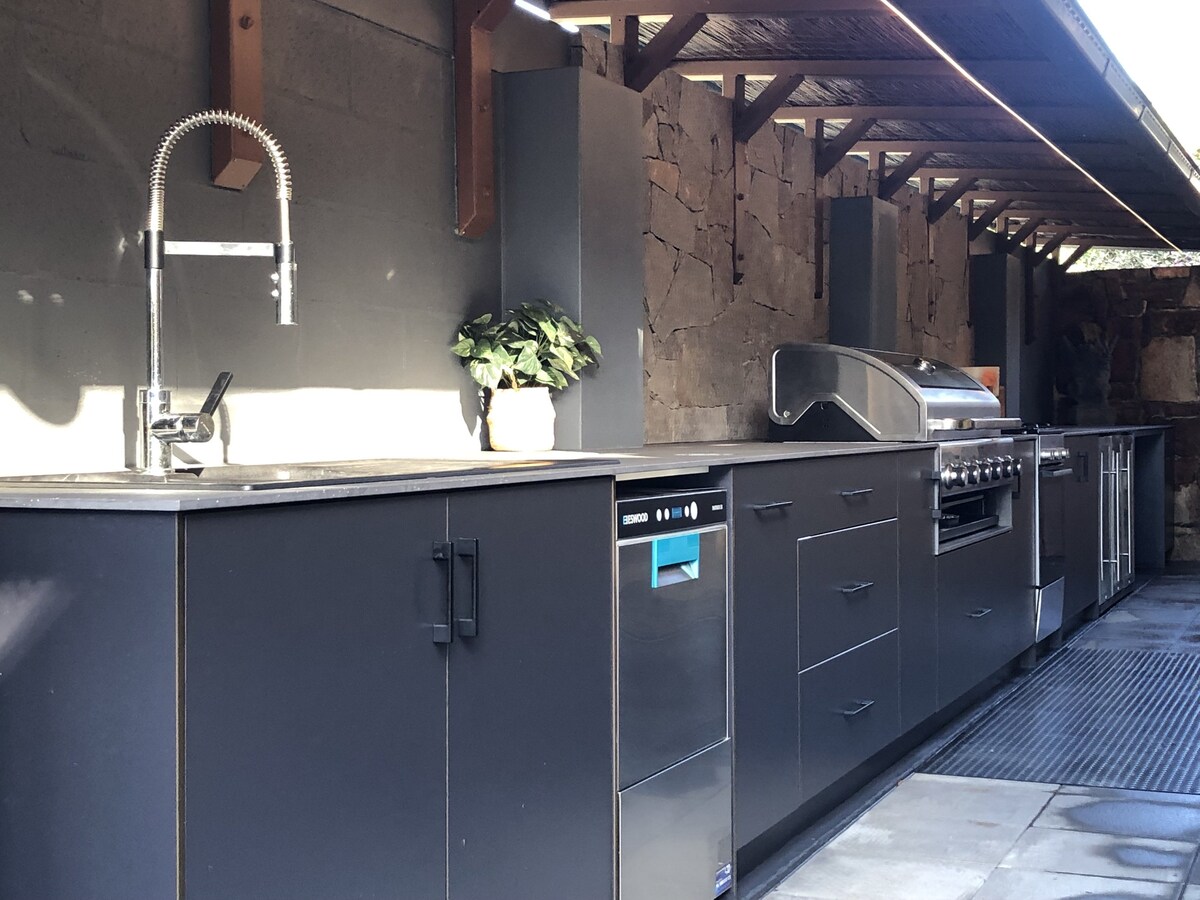 An outdoor kitchen is showcased with sleek black cabinetry and modern appliances. A built-in stone BBQ is positioned next to a dishwasher and a sink with a polished faucet. Natural light illuminates the area, enhancing its spacious layout and functionality.