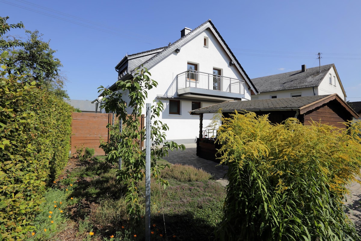 The exterior view of the modern vacation home showcases a two-story structure with a balcony. A spacious garden area surrounds the property, featuring lush greenery and a wooden shed. The clear blue sky creates a bright backdrop for this welcoming retreat.