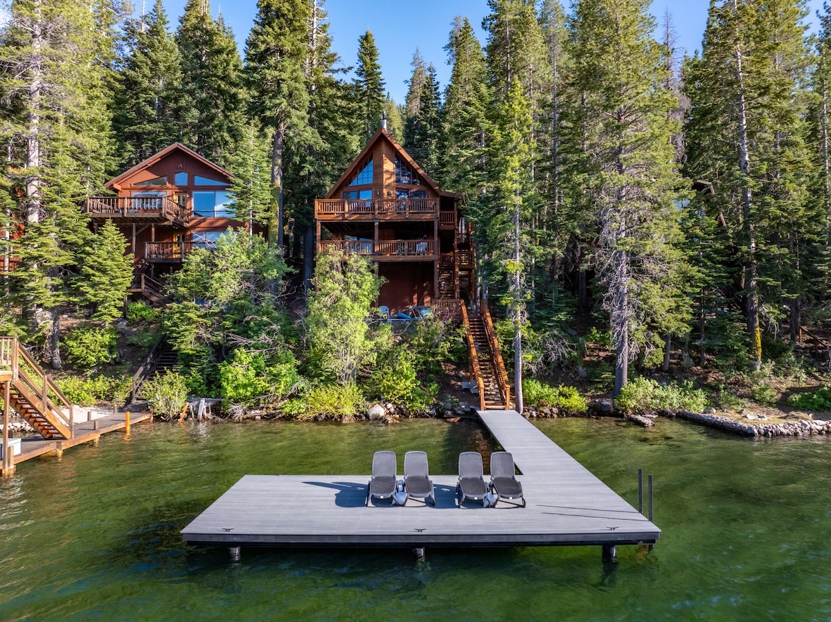 A private pier extends into the clear water, with several lounge chairs positioned for relaxation. Surrounded by tall greenery, two cabin structures are visible on the hillside, complementing the natural landscape. Stairs provide access from the pier to the cabins.