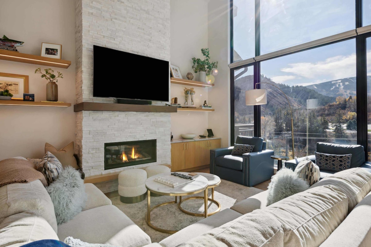 A cozy living area showcases a gas fireplace beneath a mounted television. Large windows frame panoramic views of the mountains and outdoor landscape. The plush seating includes a sectional sofa adorned with decorative pillows and a lounge chair, complemented by a round coffee table.