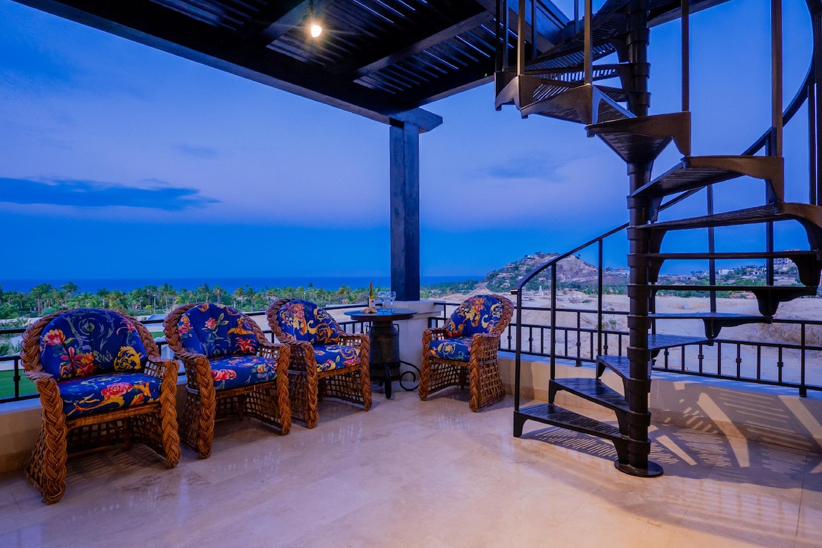 A spiral staircase leads to a cozy terrace with four woven chairs adorned in vibrant floral patterns. A small table is positioned nearby, offering an ideal spot to enjoy ocean views during twilight. Soft lighting illuminates the space, enhancing its inviting atmosphere.